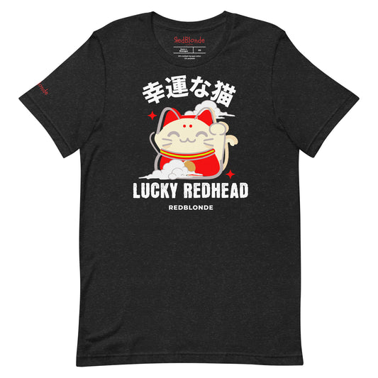 A black heather unisex T-shirt featuring a colorful maneki-neko (lucky cat) graphic wearing a red outfit with gold accents. Above the cat, Japanese kanji characters are printed in bold white. Below the illustration, the text reads “LUCKY REDHEAD” in distressed white font, with “REDBLONDE” in smaller uppercase letters underneath. The shirt has short sleeves, a classic crew neckline, and a casual, modern fit.