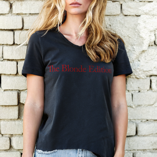 Blonde model, by a wall, wearing jeans and a black unisex relaxed fit V-neck t-shirt with “Dirty Blonde” written in vintage Coca-Cola milk-store script in red on the front