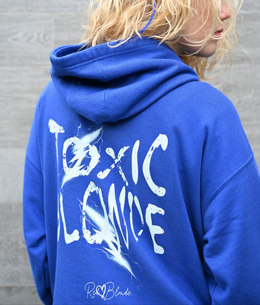 Blonde wears a royal blue unisex hoodie featuring seen from behind. It has a bold distressed white Chainsaw Carnage text reading “Toxic Blonde” with lightning-style accents and the RedBlonde signature logo underneath. The design has an edgy, electric aesthetic.It features the text 'Blonde' embroidered on the front. Branded RedBlonde in red on right sleeve.