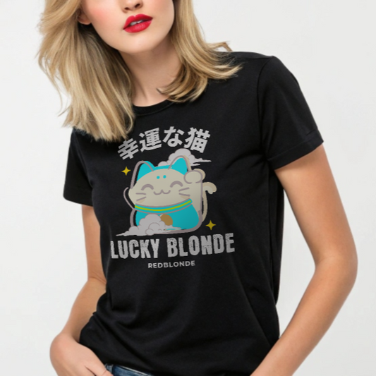 Blonde model wears a black unisex T-shirt featuring a cheerful maneki-neko (lucky cat) graphic in cream, turquoise, and black, with golden star accents. Above the cat are Japanese characters meaning “Lucky Cat.” Below the design, the text reads “LUCKY BLONDE” in bold black letters and “REDBLONDE” in smaller font. The T-shirt has a classic crew neckline and short sleeves. Branded RedBlonde on the inside neck and right sleeve