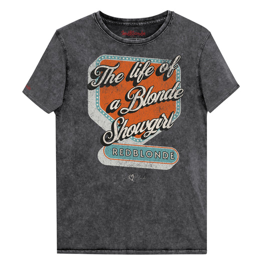 Vintage black acid-wash unisex t-shirt with bold retro-style graphic text reading 'The life of a Blonde Showgirl' in white script over an orange and turquoise marquee design. The word 'RedBlonde' is displayed in turquoise block letters at the bottom, with subtle worn details giving the shirt a distressed, retro look." Branded redblonde on inside collar and right sleeve