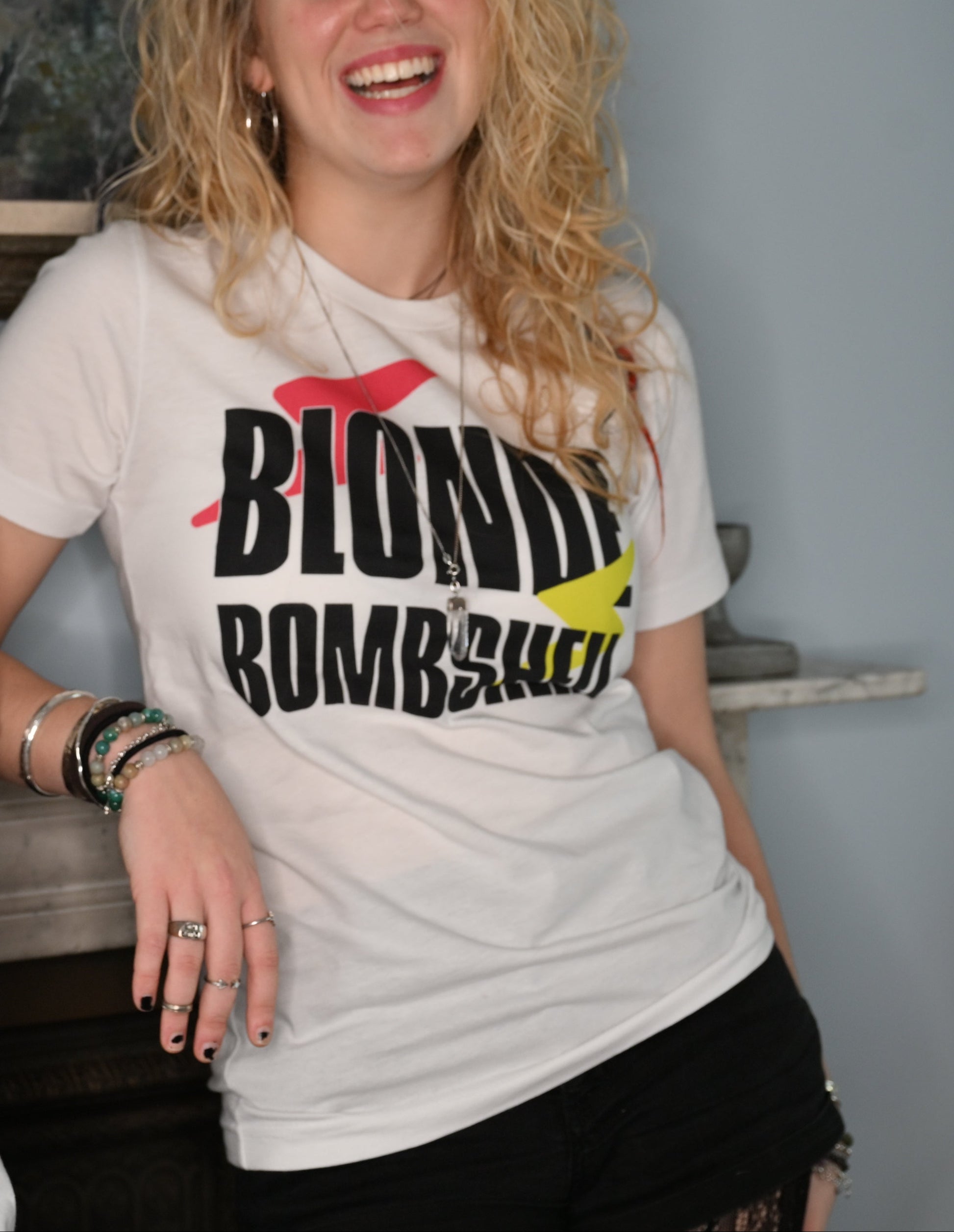 Blonde model by a wall wears a relaxed-fit white t-shirt with bold 80s ‘David Bowie’ inspired lightning graphic flashes of pink and yellow around the words ‘blonde bombshell’.