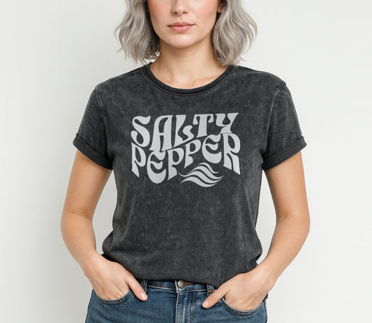 Gray-haired woman wears a soft black denim wash vintage  graphic t-shirt with the slogan 'Salty Pepper' in white retro 70s wavy font. Branded RedBlonde in red on inside neck and right sleeve