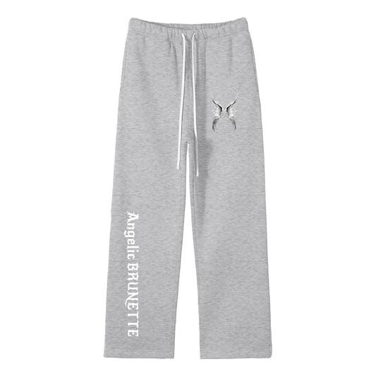 grey straight-leg joggers with white drawstrings, featuring a small white angel-wing graphic on the upper left thigh and ‘Angelic Brunette’ printed vertically down the right leg in New Rocker gothic style lettering, shown flat against a white background.