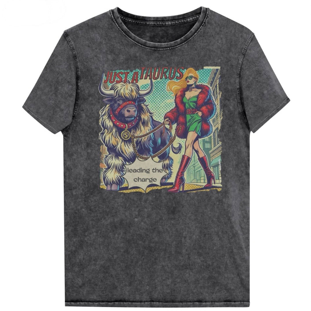 Flatlay of a unisex relaxed fit black vintage denim wash t-shirt with a retro pop-art taurus graphic featuring a blonde woman with long flowing straight hair, wearing a green dress with a red belt, red high-heeled boots and red fur coat, walking a furry bull with a red collar and medallion in city street captioned ‘Just a taurus leading the charge'. Branded RedBlonde in red on inside neck and right shoulder