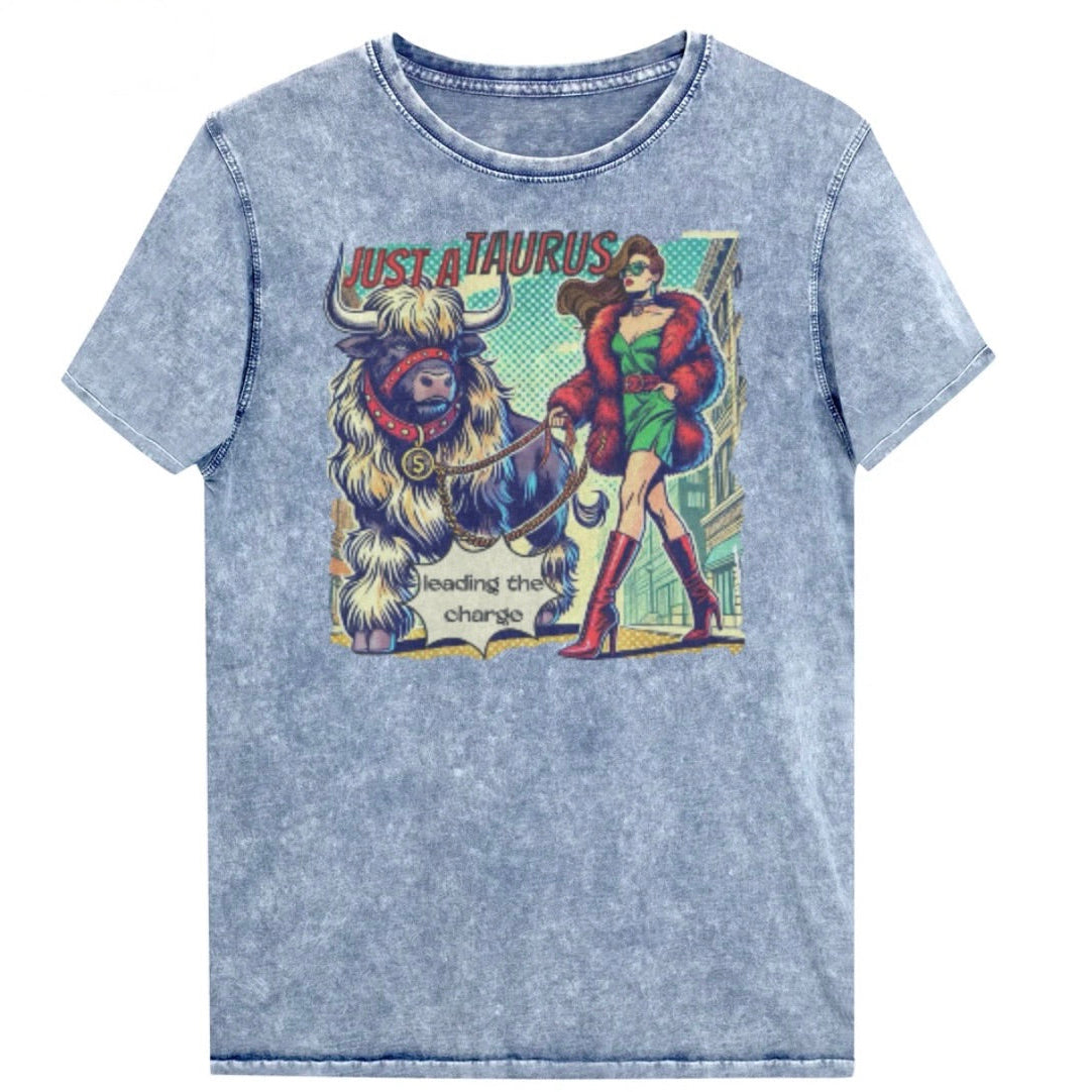 Flatlay of a unisex relaxed fit blue vintage denim wash t-shirt with a retro pop-art taurus graphic featuring a brunette woman with long flowing straight hair, wearing a green dress with a red belt, red high-heeled boots and red fur coat, walking a furry bull with a red collar and medallion in city street captioned ‘Just a taurus leading the charge'. Branded RedBlonde on inside neck and right shoulder