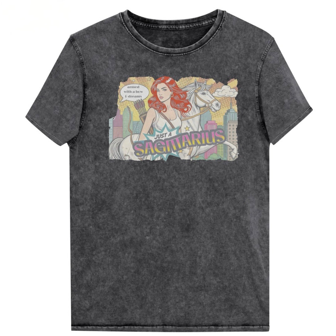 Flatlay of a unisex black denim wash vintage t-shirt with a retro pop-art Redhead Sagittarius graphic featuring a blonde woman, with bow & arrows, white stallion, and a city backdrop, captioned ‘Just a Sagittarius, armed with a bow & dreams'. Branded RedBlonde on the inside neck and right sleeve