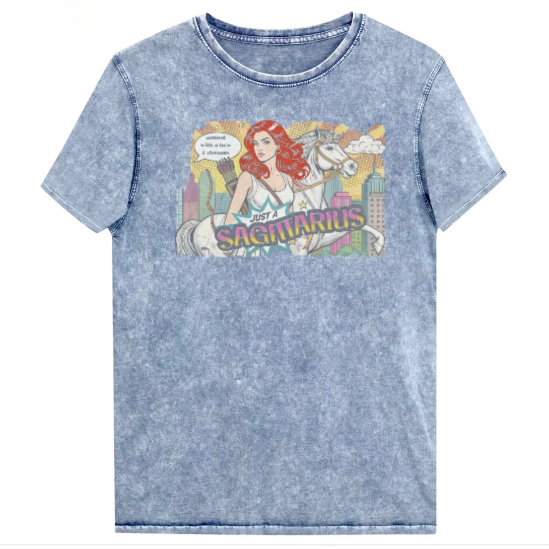 Flatlay of a unisex blue denim wash vintage t-shirt with a retro pop-art Redhead Sagittarius graphic featuring a blonde woman, with bow & arrows, white stallion, and a city backdrop, captioned ‘Just a Sagittarius, armed with a bow & dreams'. Branded RedBlonde on the inside neck and right sleeve
