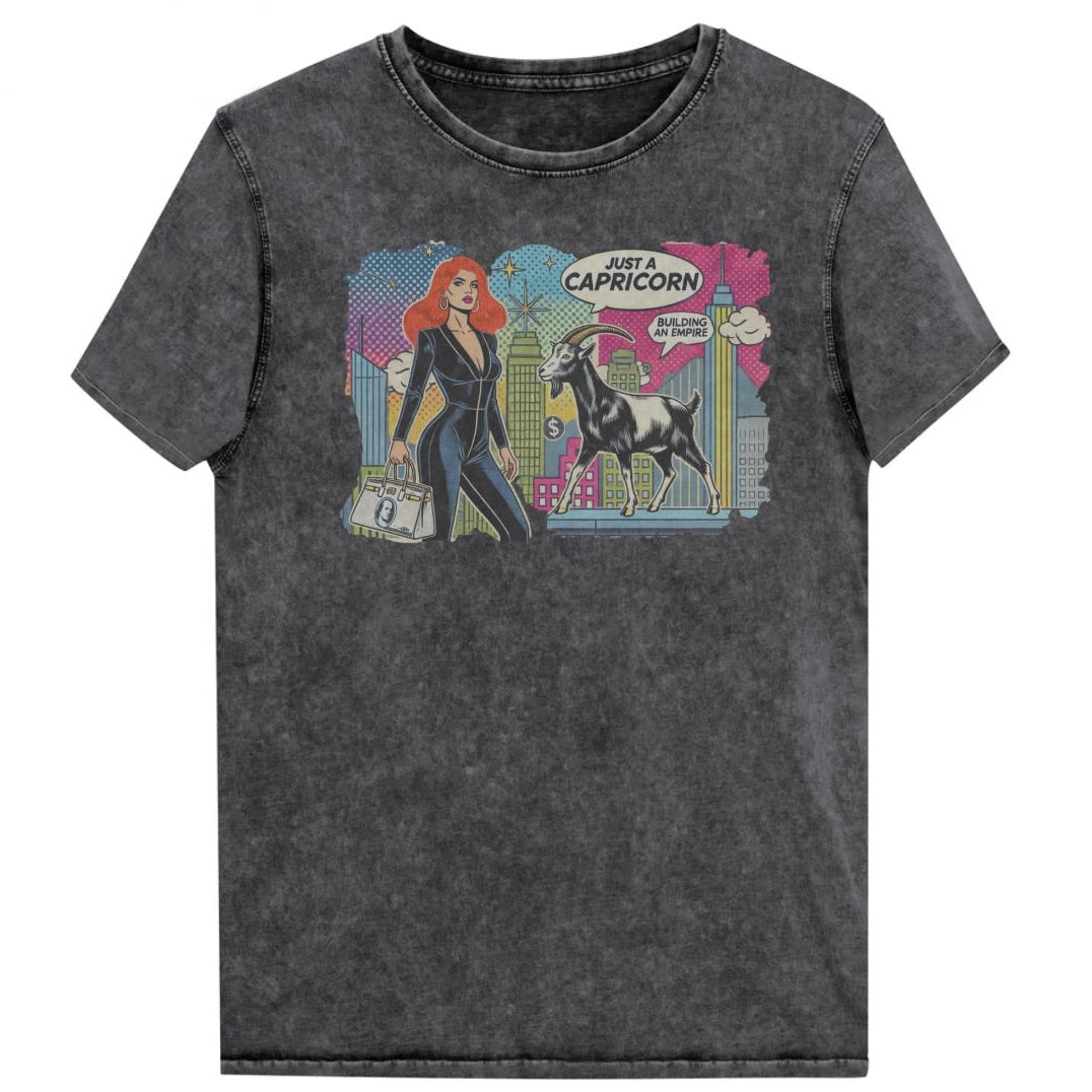 Flatlay of a unisex black denim wash vintage t-shirt with a retro pop-art Capricorn graphic featuring a red haired woman in a black catsuit, carrying a bag, with a goat  and a city skyscraper backdrop, captioned ‘Just a Capricorn, Building an empire'. Branded RedBlonde on inside neck and right sleeve
