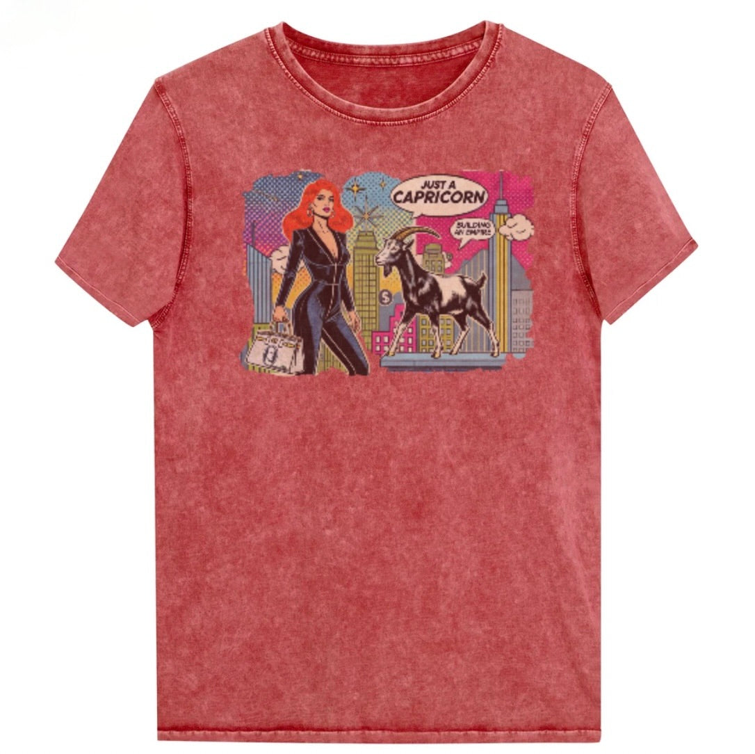 Flatlay of a unisex red garnet denim wash vintage t-shirt with a retro pop-art Capricorn graphic featuring a red haired woman in a black catsuit, carrying a bag, with a goat  and a city skyscraper backdrop, captioned ‘Just a Capricorn, Building an empire'. Branded RedBlonde on inside neck and right sleeve