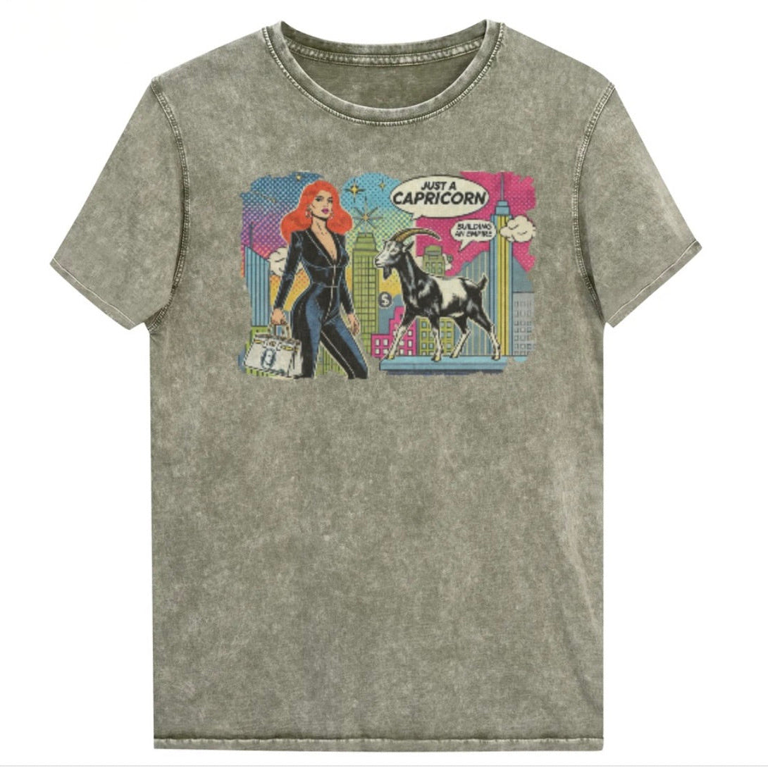 Flatlay of a unisex army green denim wash vintage t-shirt with a retro pop-art Capricorn graphic featuring a red haired woman in a black catsuit, carrying a bag, with a goat  and a city skyscraper backdrop, captioned ‘Just a Capricorn, Building an empire'. Branded RedBlonde on inside neck and right sleeve
