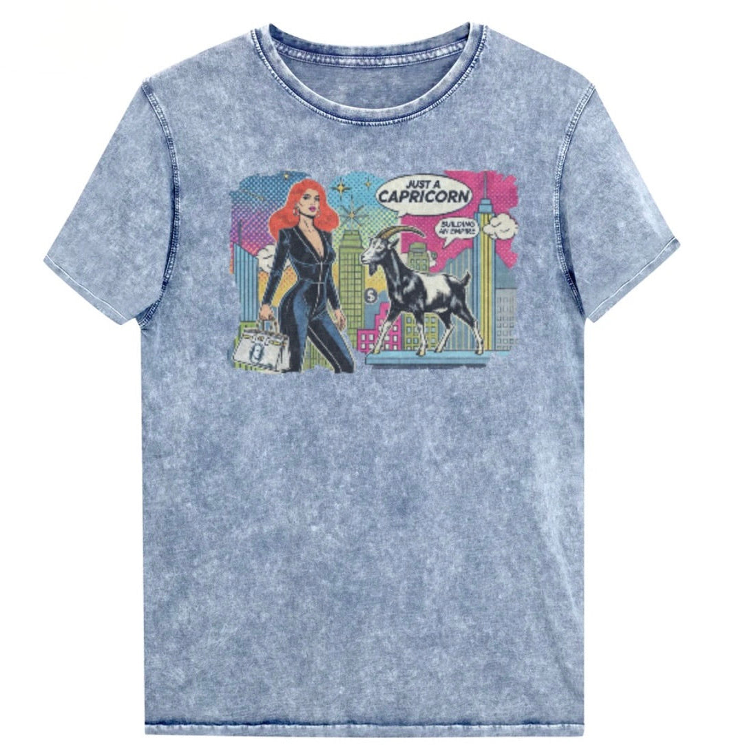 Flatlay of a unisex blue denim wash vintage t-shirt with a retro pop-art Capricorn graphic featuring a red haired woman in a black catsuit, carrying a bag, with a goat  and a city skyscraper backdrop, captioned ‘Just a Capricorn, Building an empire'. Branded RedBlonde on inside neck and right sleeve