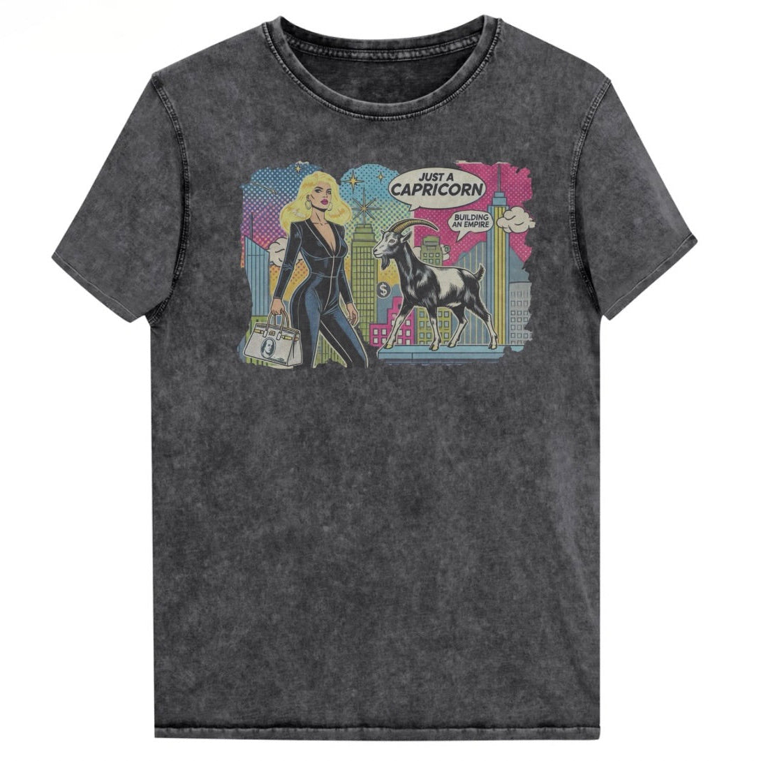 Flatlay of a unisex black denim wash vintage t-shirt with a retro pop-art Capricorn graphic featuring a blonde haired woman in a black catsuit, carrying a bag, with a goat  and a city skyscraper backdrop, captioned ‘Just a Capricorn, Building an empire'. Branded RedBlonde in red on inside neck and right sleeve