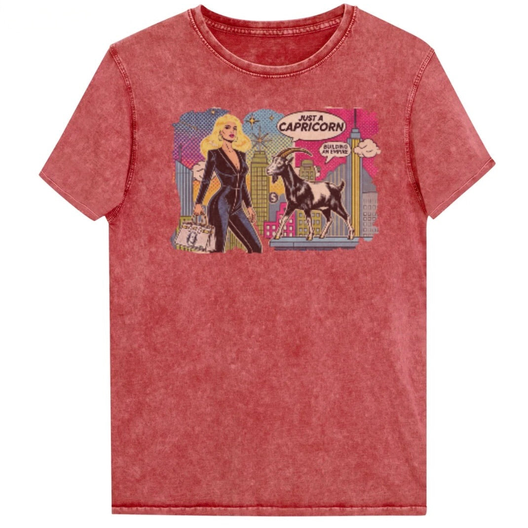 Flatlay of a unisex garnet red denim wash vintage t-shirt with a retro pop-art Capricorn graphic featuring a blonde haired woman in a black catsuit, carrying a bag, with a goat  and a city skyscraper backdrop, captioned ‘Just a Capricorn, Building an empire'. Branded RedBlonde in red on inside neck and right sleeve