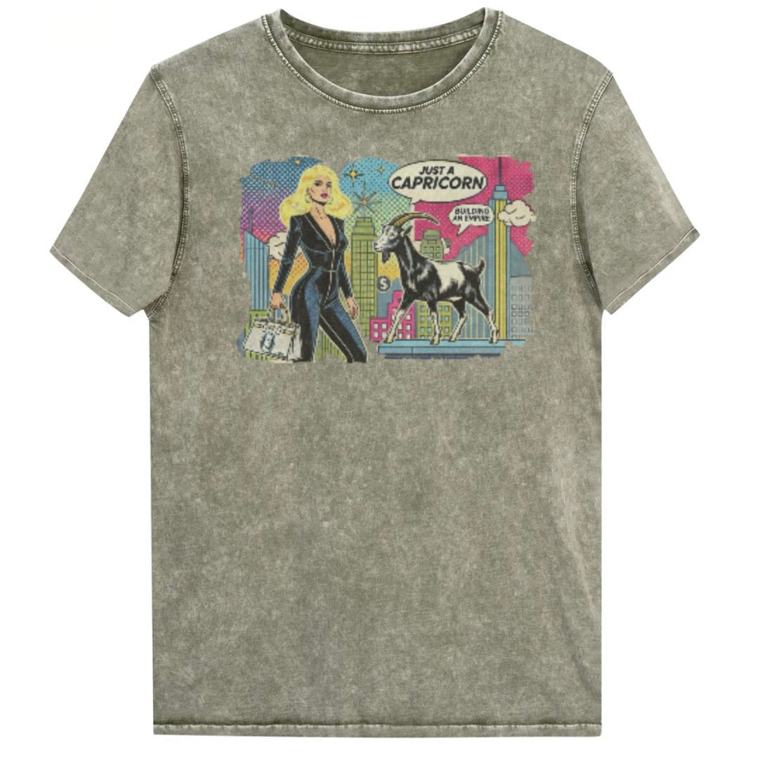 Flatlay of a unisex army green denim wash vintage t-shirt with a retro pop-art Blonde Capricorn graphic featuring a blonde haired woman in a black catsuit, carrying a bag, with a goat  and a city skyscraper backdrop, captioned ‘Just a Capricorn, Building an empire'. Branded RedBlonde in red on inside neck and right sleeve