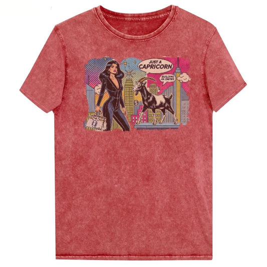 Flatlay of a unisex garnet red denim wash vintage t-shirt with a retro pop-art Capricorn graphic featuring a black haired woman in a black catsuit, carrying a bag, with a goat  and a city skyscraper backdrop, captioned ‘Just a Capricorn, Building an empire'. Branded RedBlonde on inside neck and right sleeve