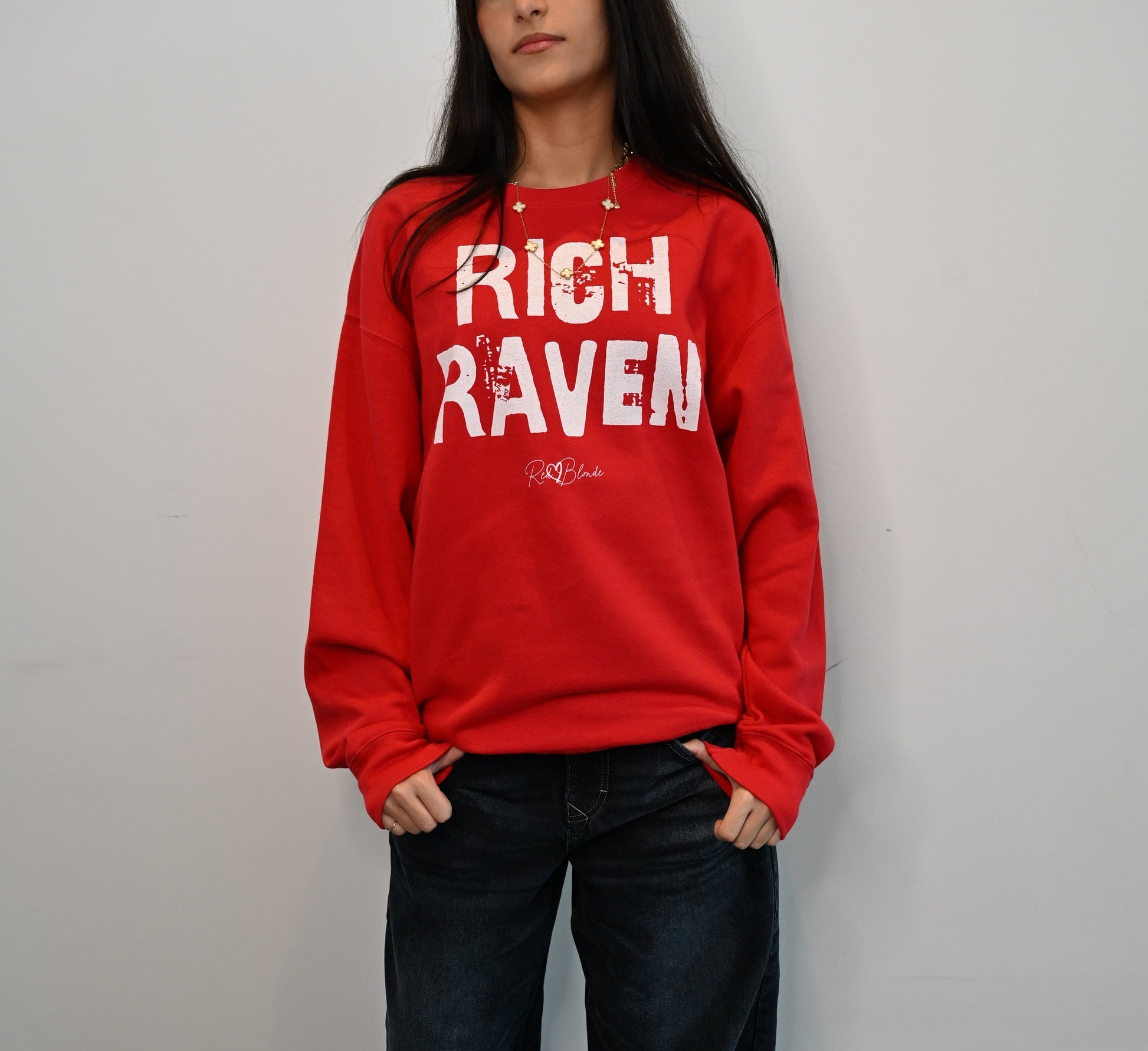 Model with jet black hair wears a White unisex premium sweatshirt with bold distressed Red Ed Geinitext “RICH RAVEN” on the front, featuring a subtle RedBlonde logo beneath the lettering and on the sleeve cuff and inside neck