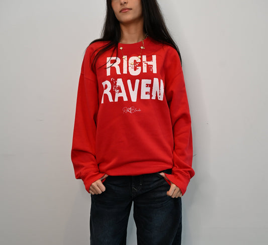 Model with jet black hair wears a White unisex premium sweatshirt with bold distressed Red Ed Geinitext “RICH RAVEN” on the front, featuring a subtle RedBlonde logo beneath the lettering and on the sleeve cuff and inside neck
