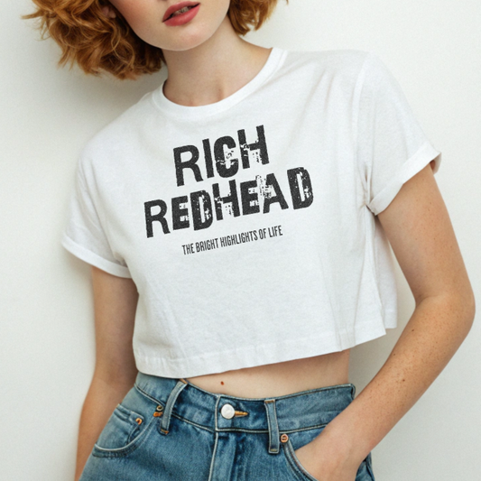 Redhead wears A white women’s crop top with a relaxed fit, short sleeves, and a wide crew neckline. The front features bold black distressed lettering reading “RICH REDHEAD” with the tagline “THE BRIGHT HIGHLIGHTS OF LIFE” in smaller text below. 