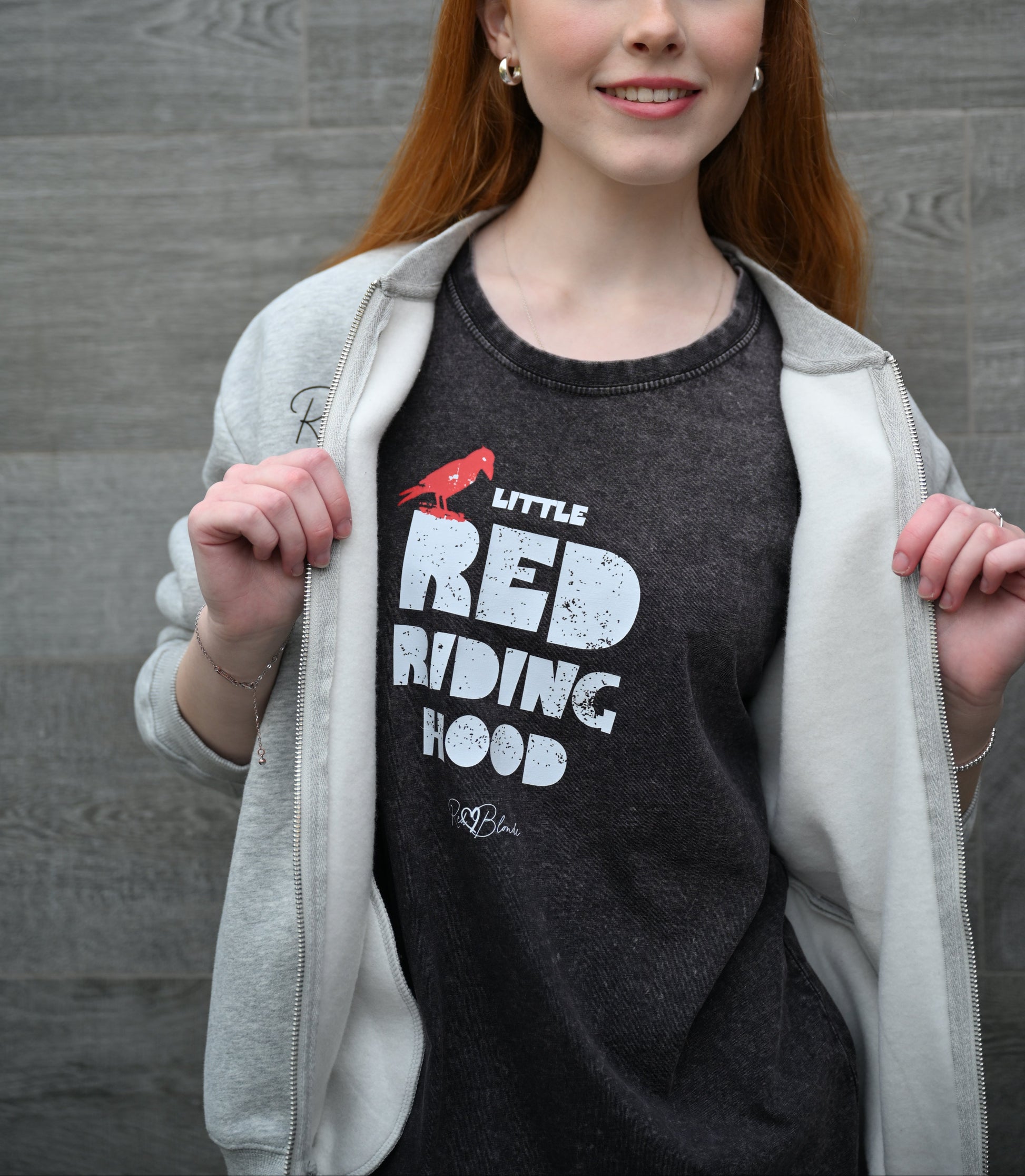 Model wears black vintage acid wash denim look unisex t-shirt featuring bold distressed Love Birds font in white reading “Little Red Riding Hood” with a small red lack crow perched on the word “Little,” and the RedBlonde signature logo below. Branded RedBlonde London on inside neck and right sleeve