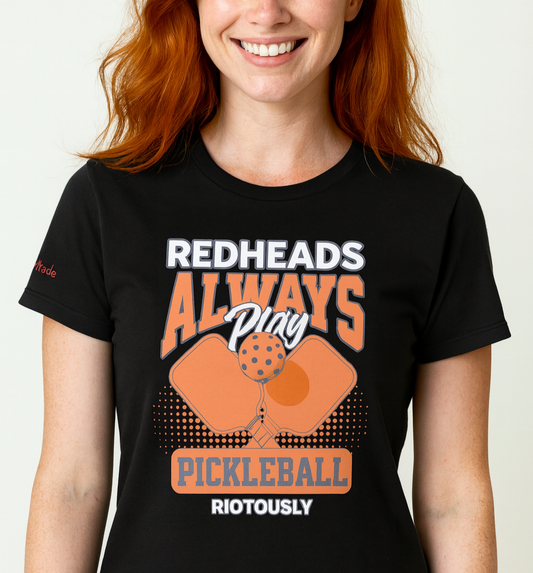 Redhead wears a Black unisex t-shirt with orange graphic that reads "Redheads Always Play Pickleball Riotously," featuring pickleball paddles and ball. Branded RedBlonde on inside neck and right sleeve