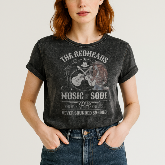 Redhead wears a black denim wash unisex t-shirt with ‘THE REDHEADS’, ‘MUSIC FROM THE SOUL’, ‘RED HAIR AND RED LIPS’, and ‘NEVER SOUNDED SO GOOD’ written in Sancreek font features a graphic of a redheaded cowgirl singing with two guitars behind her. Branded RedBlonde London in red on inside neck and right sleeve 