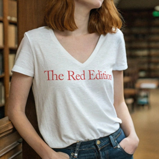 Redhead woman in library wears a slightly fitted white v-neck t-shirt with the words 'The Red Edition' in red. Branded RedBlonde in red on the inside neck and right sleeve