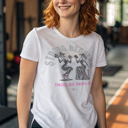 Redhead woman wears a white relaxed-fit crew neck t-shirt featuring a graphic of two women: one holding a barbell on her shoulders, the other reading a book on a pile of books. The words ‘STRONG REDHEAD’ are arced over the women in pale grey, while ‘Powerful Body, Powerful Mind’ is in bright pink underneath. The t-shirt is signed off RedBlonde with a heart. Branded RedBlonde in red on inside neck and right sleeve