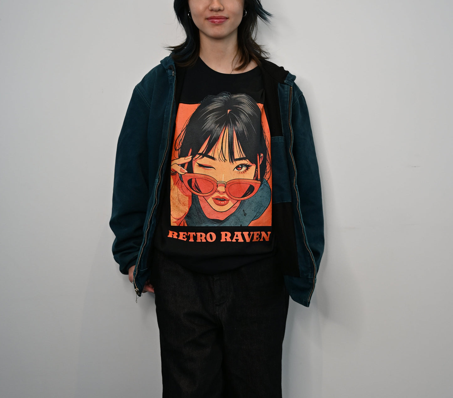 Raven haired woman wears a white unisex t-shirt featuring a vintage-style illustrated graphic of a woman with black hair winking while lowering orange sunglasses. The design is framed in warm retro tones with the words “RETRO RAVEN” printed below in bold orange text. The RedBlonde logo appears inside the neckline and subtly on the sleeve.