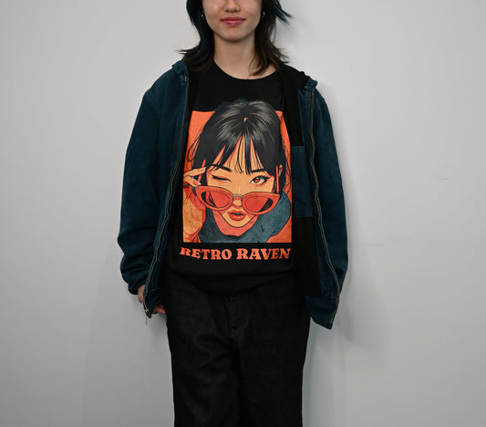 Raven haired woman wears a white unisex t-shirt featuring a vintage-style illustrated graphic of a woman with black hair winking while lowering orange sunglasses. The design is framed in warm retro tones with the words “RETRO RAVEN” printed below in bold orange text. The RedBlonde logo appears inside the neckline and subtly on the sleeve.