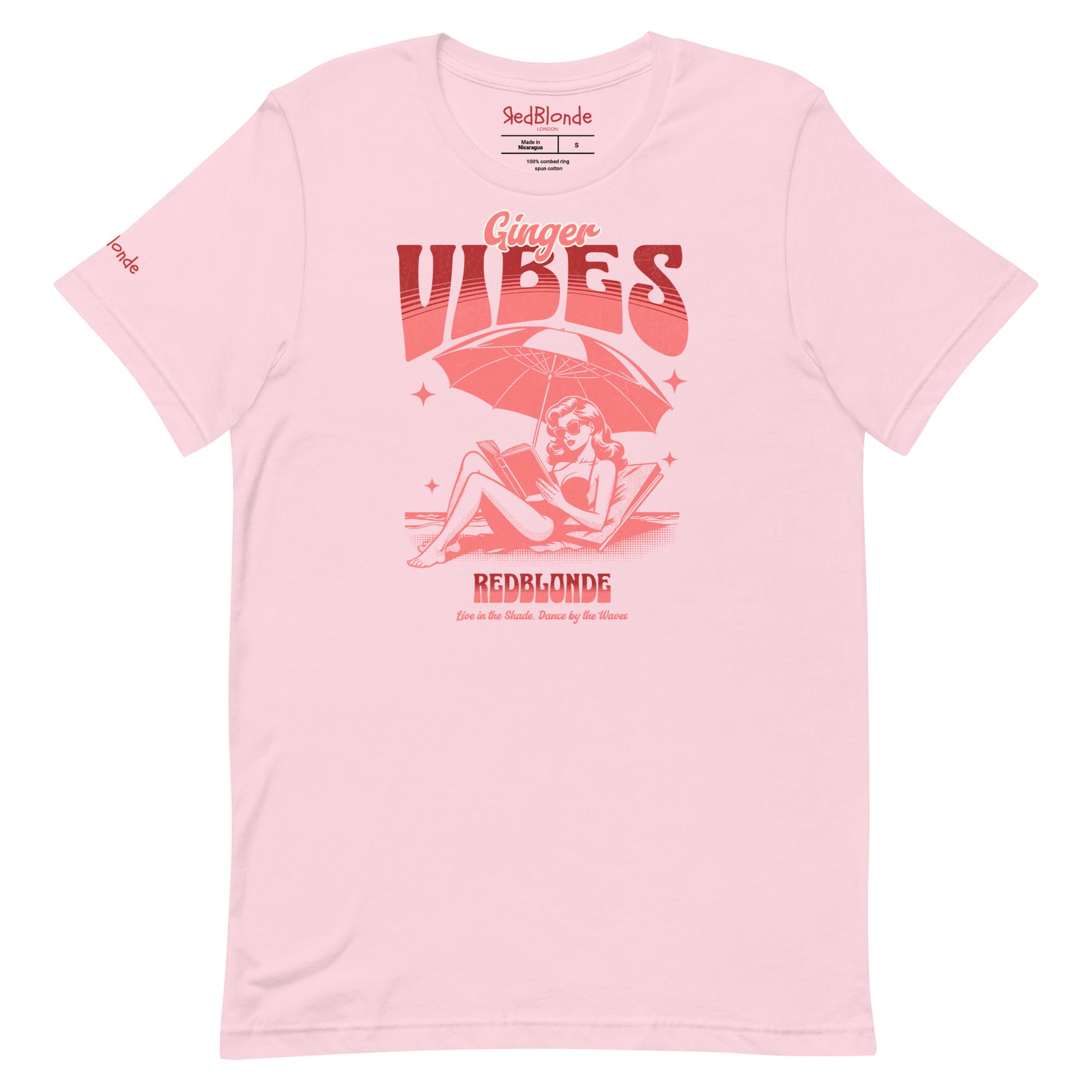 Soft pink unisex t-shirt featuring a retro-style red illustration of a redhead woman lounging under a beach umbrella reading a book, with bold text reading "Ginger Vibes" above and "RedBlonde" below, alongside the tagline "Live in the shade. Dance by the waves." Branded RedBlonde on inside neck and right sleeve
