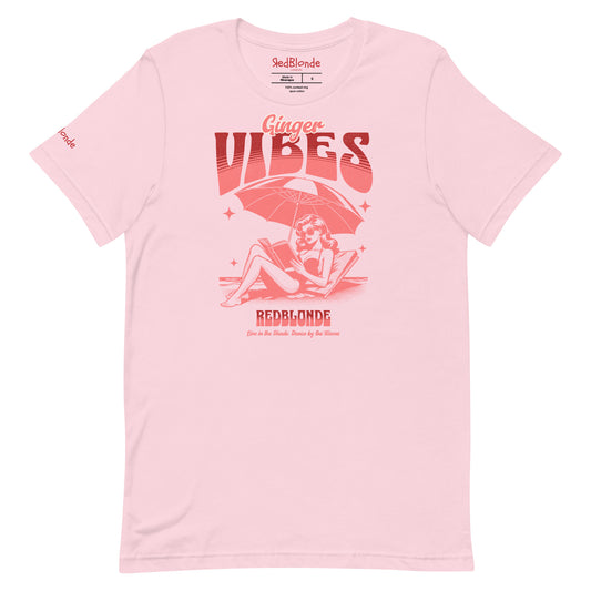 Soft pink unisex t-shirt featuring a retro-style red illustration of a redhead woman lounging under a beach umbrella reading a book, with bold text reading "Ginger Vibes" above and "RedBlonde" below, alongside the tagline "Live in the shade. Dance by the waves." Branded RedBlonde on inside neck and right sleeve