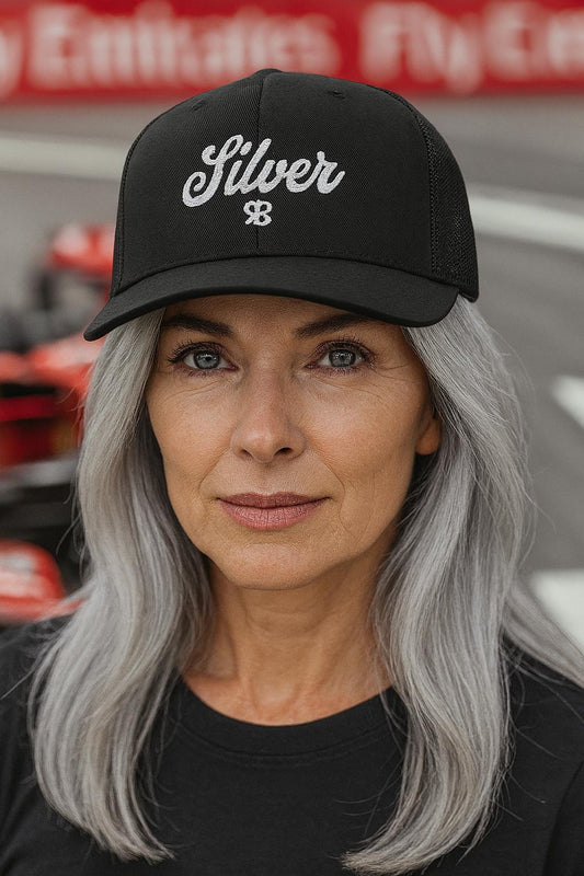 Woman with grey hair wears a black  trucker cap with 'Silver' embroidered in white adventurer 01 font on front. Mesh back. Branded RB