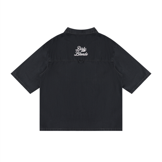 back view of a black, short-sleeve button-up shirt with a subtle faded finish and a small raised “Dirty Blonde” Milkstore script logo centered across the upper back.