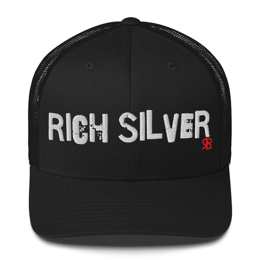 Black mesh-back trucker cap with the bold distressed text “RICH SILVER” embroidered in white across the front, and a small red “RB” logo detail beneath the text. The cap has a structured front panel and curved brim