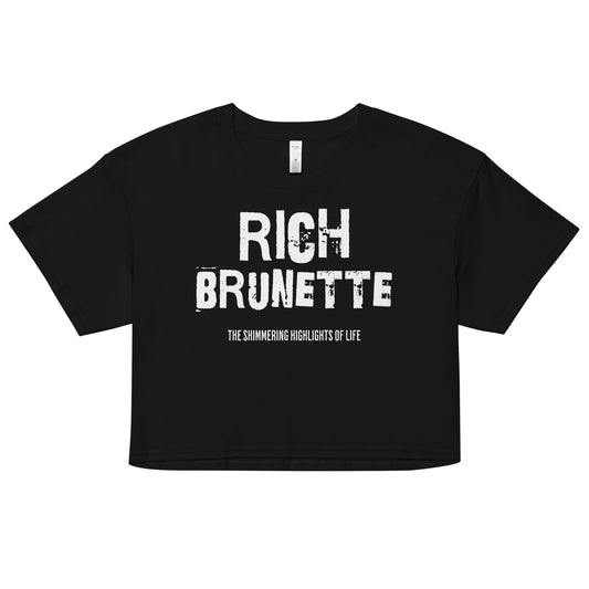 A black women’s crop top with a relaxed fit, short sleeves, and a wide crew neckline. The front features bold white distressed lettering reading “RICH BRUNETTE” with the tagline “THE SHIMMERING HIGHLIGHTS OF LIFE” in smaller text below. 