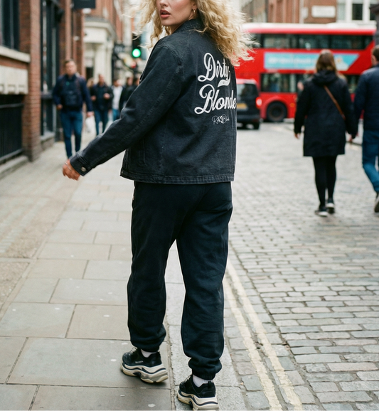 Model wears a washed-black denim jacket showing large white embroidered Miklstore font script reading "Dirty Blonde" with the RedBlonde signature below. The jacket has a relaxed, slightly cropped silhouette and long sleeves with button cuffs.