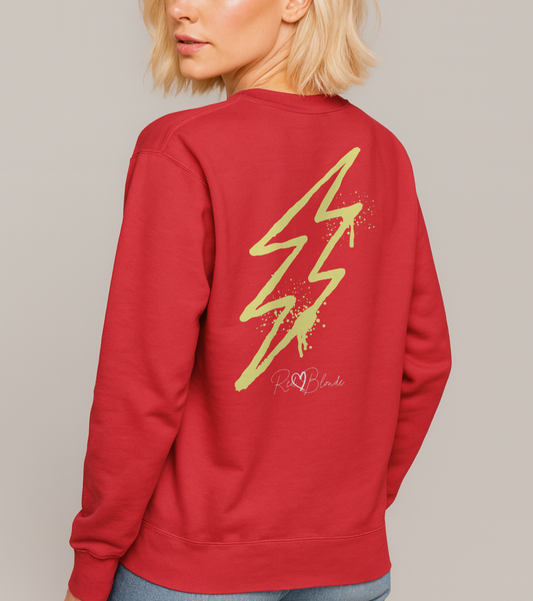 blonde wears a red unisex sweatshirt featuring a bold yellow lightning bolt graphic with paint-splatter detail and the RedBlonde logo printed below. The design has an edgy, electric streetwear vibe. Branded RedBlonde in red on right sleeve