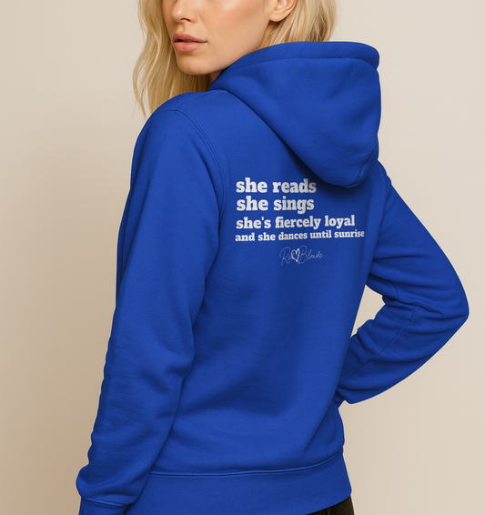 Blonde wears a royal blue unisex hoodie viewed from the back featuring white text that reads, “she reads, she sings, she’s fiercely loyal, and she dances until sunrise,” with the RedBlonde logo printed underneath. The design conveys confidence and personality with a bold, uplifting message.