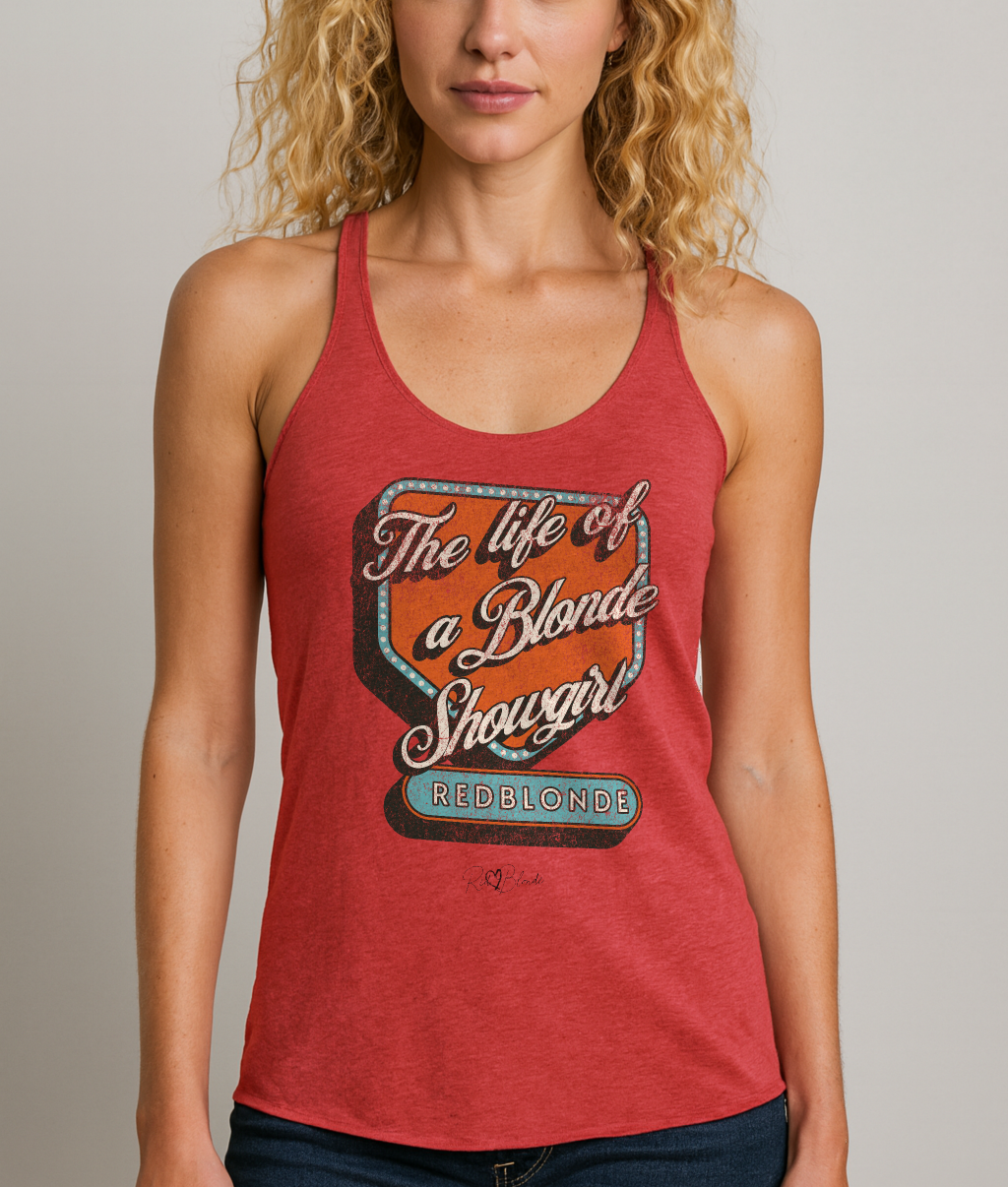 blonde wears a Vintage red racerback tank t-shirt with bold retro-style graphic text reading 'The life of a Blonde Showgirl' in white script over an orange and turquoise marquee design. The word 'RedBlonde' is displayed in turquoise block letters at the bottom, with subtle worn details giving the shirt a distressed, retro look." 