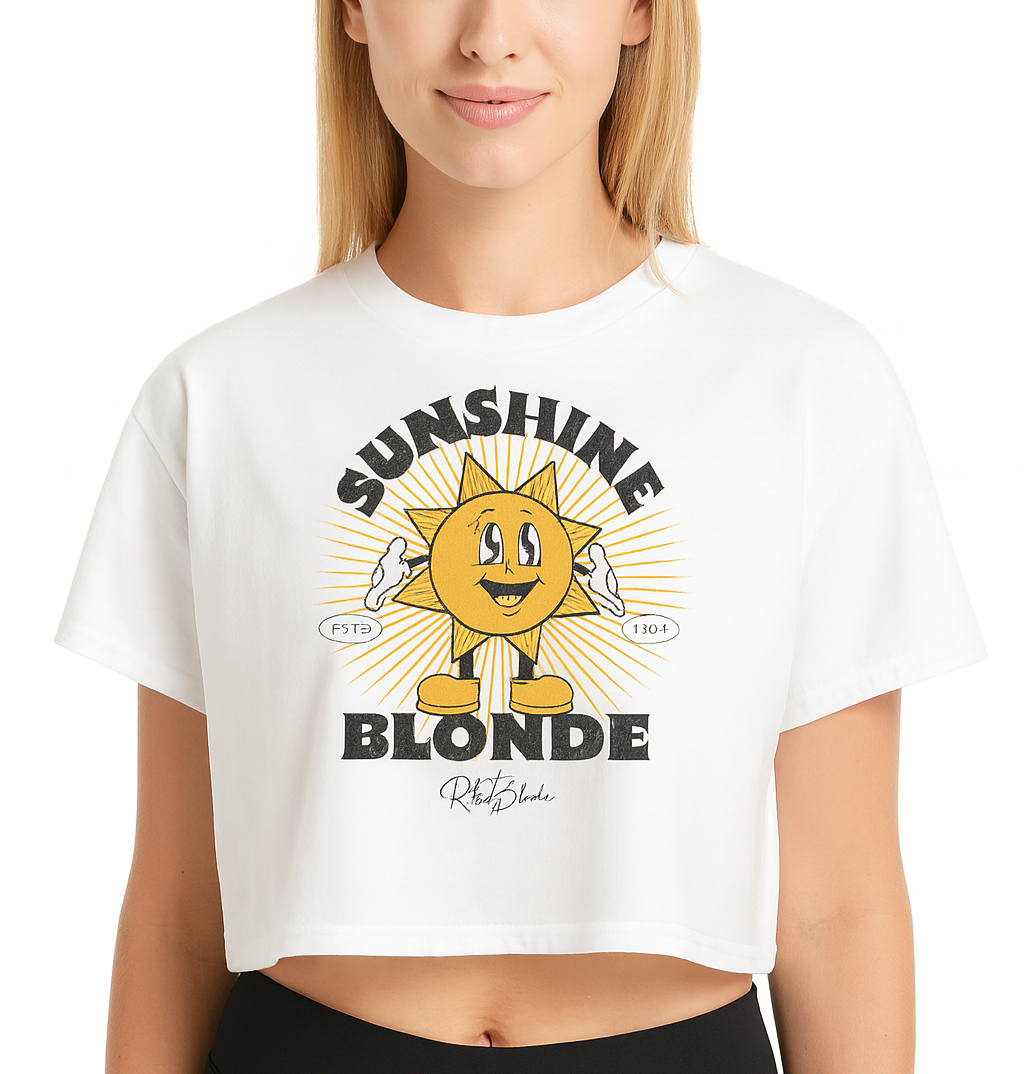 blonde woman wears a White women’s crop top with a retro cartoon-style yellow sun character, smiling with open arms, surrounded by radiant sunbeams. The text reads ‘Sunshine Blonde’ in bold vintage Belzebuth lettering, with small details including ‘ESTD 1994’ and the RedBlonde signature logo beneath."
