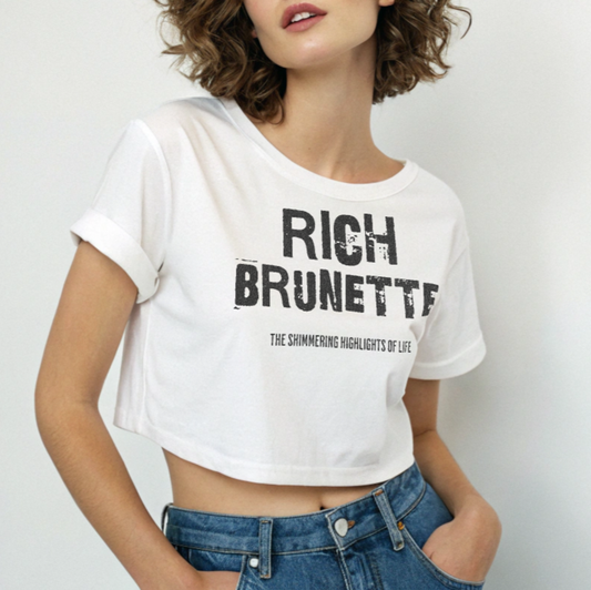 brunette wears A white women’s crop top with a relaxed fit, short sleeves, and a wide crew neckline. The front features bold black distressed lettering reading “RICH BRUNETTE” with the tagline “THE SHIMMERING HIGHLIGHTS OF LIFE” in smaller text below. 