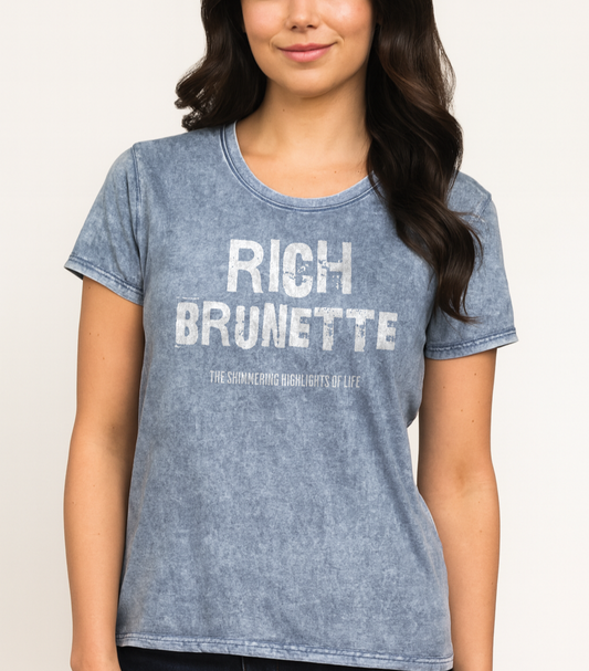 Brunette wears a A blue acid-wash unisex denim T-shirt with a distressed vintage look. The shirt features the bold white text “RICH BRUNETTE” in a weathered Ed Gein font, with the tagline “THE SHIMMING HIGHLIGHTS OF LIFE” in smaller uppercase letters below. The shirt has short sleeves, a crew neckline, and visible seam detailing for a rugged, casual style. Branded Redblonde on inside neck and right sleeve