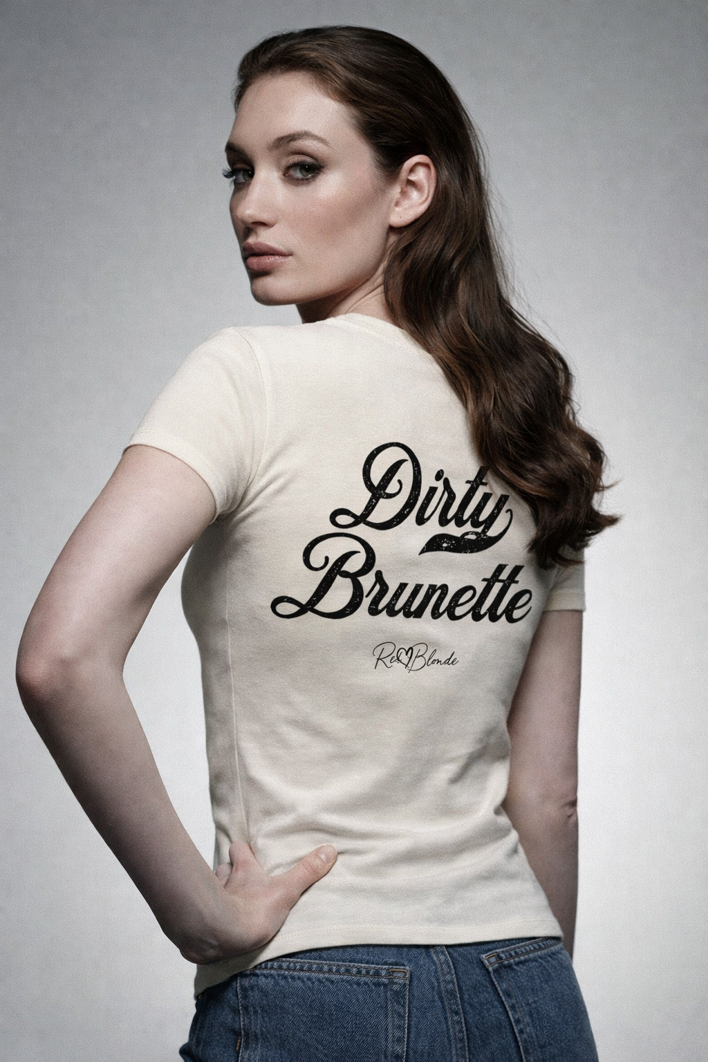 Brunette wears a fitted, short-sleeve off white T-shirt laid flat on a white background. It features large black Milkstore 01 script text reading “Dirty Brunette” across the upper back, with a small cursive “RedBlonde” logo beneath. The hem is slightly curved.