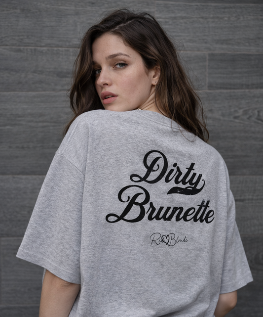 Brunette wears a light grey oversized boxy short-sleeve T-shirt laid flat on a white background. Large distressed black Milkstore 01 script text reads “Dirty Brunette,” with a small cursive “RedBlonde” logo beneath the main graphic.