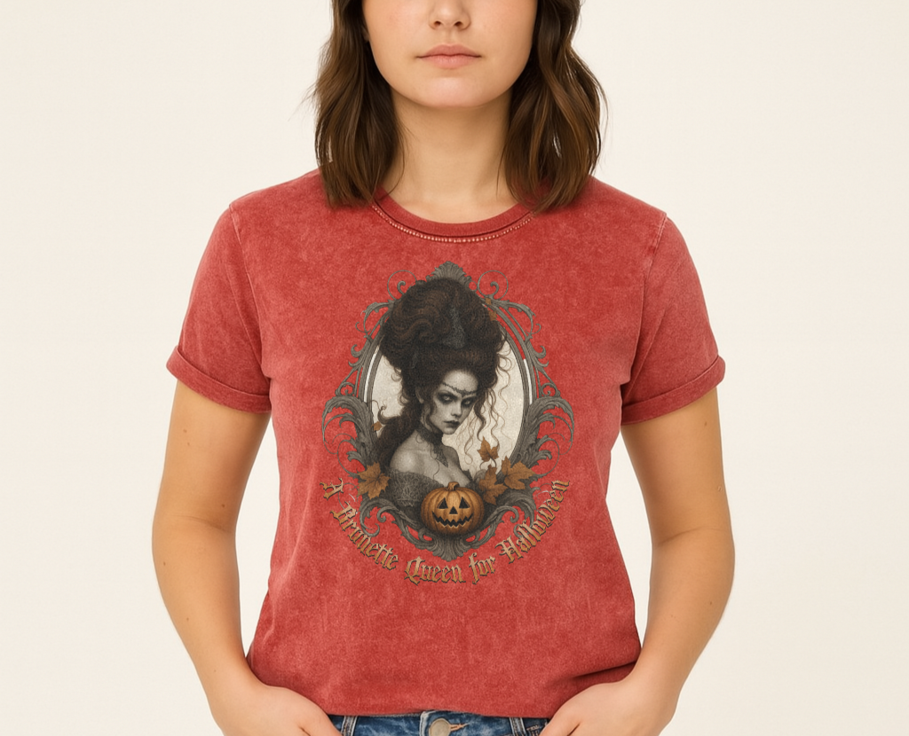 brunette wears red garnet acid-wash unisex T-shirt featuring a gothic-style illustration of a dark-haired woman with an elaborate beehive hairstyle, framed by ornate scrollwork, autumn leaves, and a carved pumpkin. The design is captioned with the text ‘A Brunette Queen for Halloween’ in vintage Dark Angle lettering. Branded RedBlonde in red on inside neck and right sleeve
