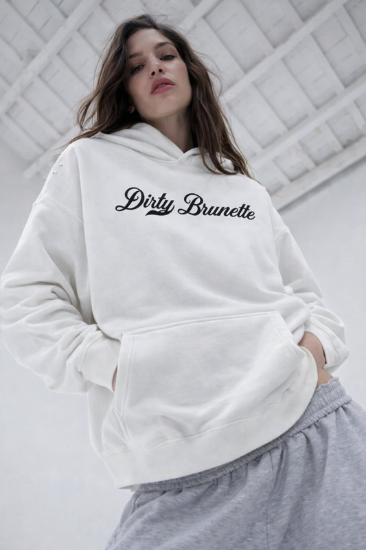 A brunette wears white pullover hoodie laid flat on a white background. It has a kangaroo front pocket and ribbed cuffs and hem. Across the chest is white Milkstore 01 script text reading “Dirty Brunette.”