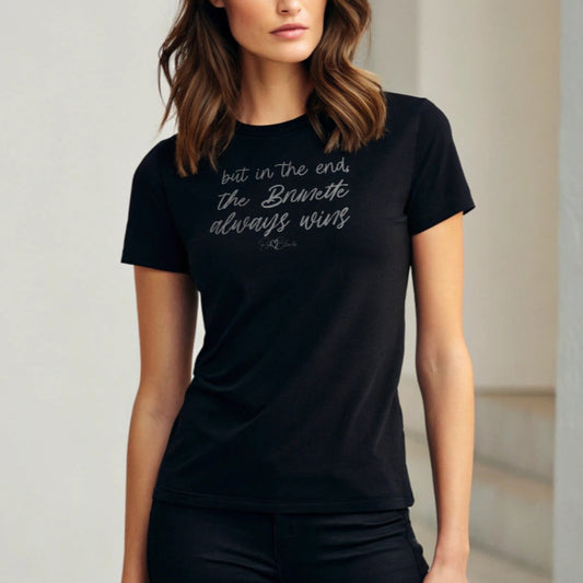 brunette woman wears black unisex t-shirt featuring the handwritten-style slogan “but in the end, the Brunette always wins” in Hillbear font  in light gray text across the chest, signed off RedBlonde. Branded “RedBlonde London on inside the collar and on the right sleeve