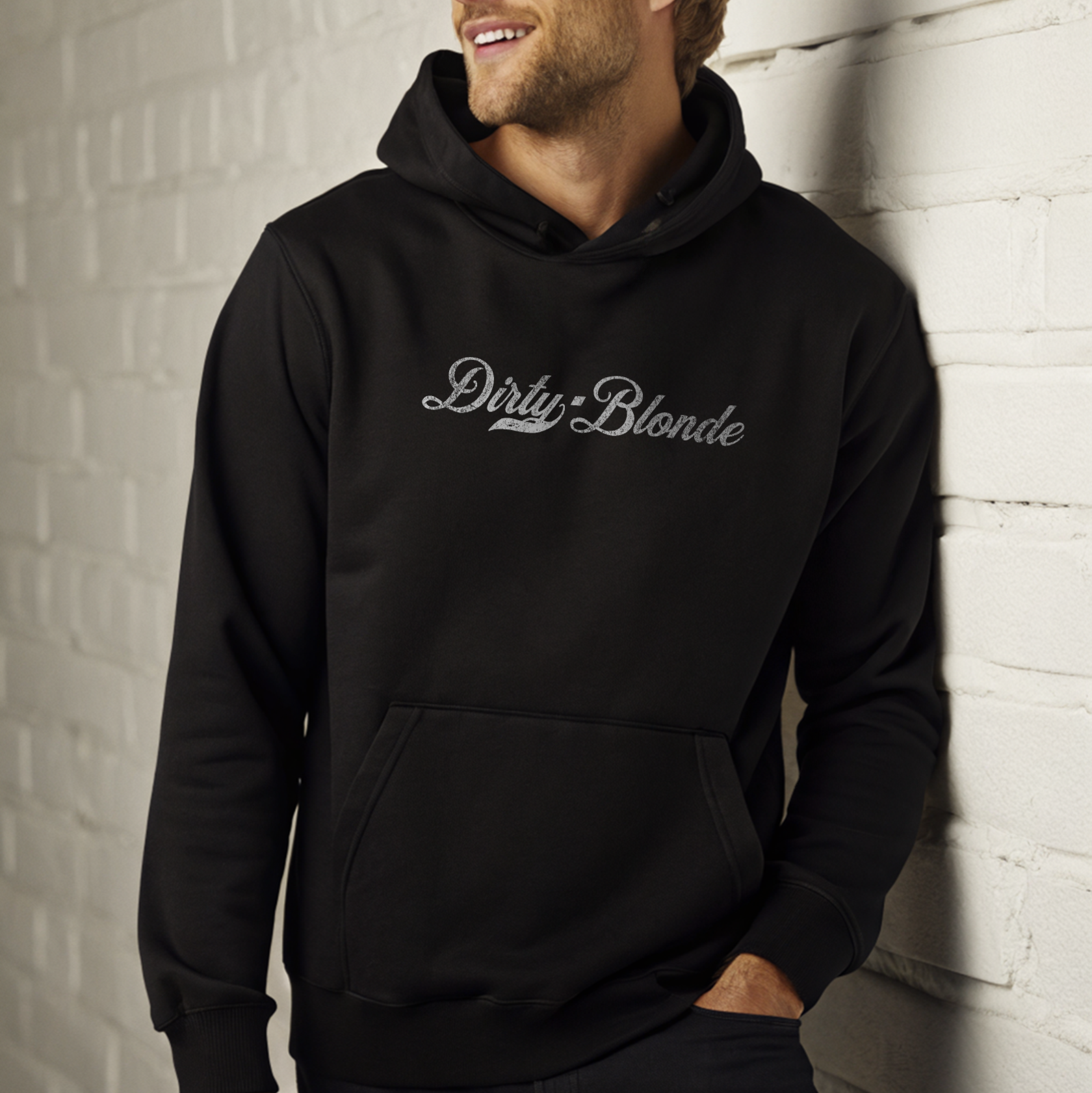 Blonde smiling man leaning on a white wall wearing a classic black relaxed fit unisex hoodie with the words 'dirty Blonde' written in white vintage milkstore font, like coco cola script 