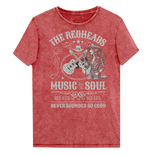 A garnet red denim wash unisex t-shirt with ‘THE REDHEADS’, ‘MUSIC FROM THE SOUL’, ‘RED HAIR AND RED LIPS’, and ‘NEVER SOUNDED SO GOOD’ written in Sancreek font features a graphic of a redheaded cowgirl singing with two guitars behind her. Branded RedBlonde London in red on inside neck and right sleeve 